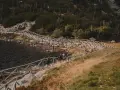 Dwie osoby idą kamiennym szlakiem przy jeziorze, otoczonym drewnianym ogrodzeniem. W oddali widać las i góry.