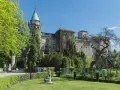 Zamek na szczytniku, otoczony zielenią, z wieżą zwieńczoną stożkowym dachem. W oddali widać bramę wjazdową.