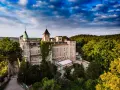 Szczytna zamek, otoczony zielenią, z wieżami i zielonym dachem, pod błękitnym niebem z chmurami.