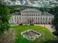 Pałac Jedlinka, biała, okazała budowla z szarym dachem, otoczona zielenią i górami. Przed pałacem fontanna i parking z samochodami.