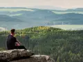 Mężczyzna siedzi na skale, podziwiając rozległy krajobraz gór i lasów. Widok przypomina skalne miasto, gdzie drzewa tworzą gęste, zielone połacie.