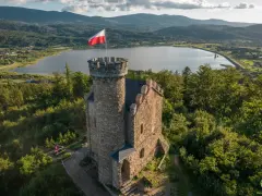 Kamienny zamek henryka z polską flagą na wieży, otoczony lasem, z jeziorem i górami w tle.