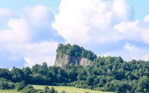 Skalisty szczyt porośnięty drzewami, przypominający wilczą górę, góruje nad zielonymi polami i lasami.