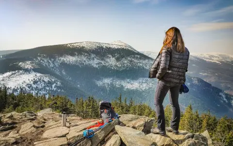 Kobieta na skalnym stole podziwia zimowy krajobraz gór. Obok niej plecak i kijki trekkingowe.