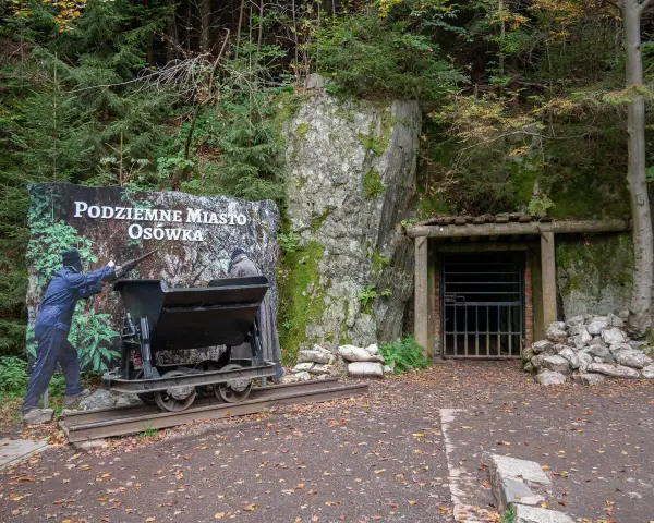Wejście do Podziemnego Miasta Osówka, z wagonikiem i tablicą informacyjną. Widać skalne ściany i wejście do tunelu.