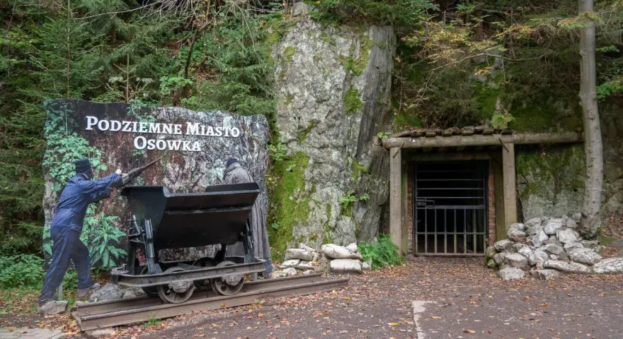 Wejście do Podziemnego Miasta Osówka, z wagonikiem i tablicą informacyjną. Widać skalne ściany i wejście do tunelu.