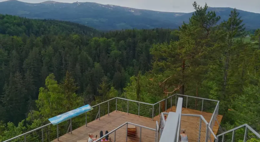 Drewniany punkt widokowy w Karpaczu z widokiem na góry i las. Dwie osoby odpoczywają na tarasie.