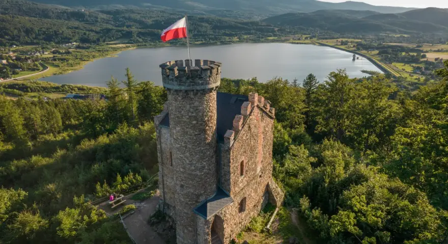 Kamienny zamek księcia Henryka z polską flagą na wieży, otoczony lasem, z widokiem na jezioro i góry. Punkt widokowy zaprasza do podziwiania krajobrazu.