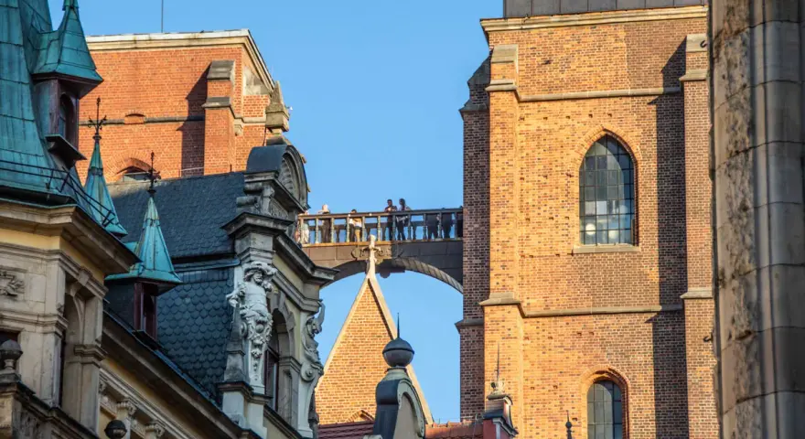 Most zabytkowych budynków we Wrocławiu z ludźmi na moście i wieżą Katedry w tle.