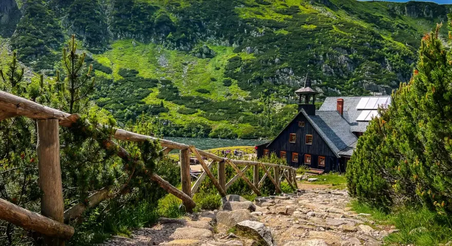 Kamienna ścieżka prowadzi do schroniska Samotnia Śnieżka nad jeziorem, otoczonego zielonymi zboczami gór.
