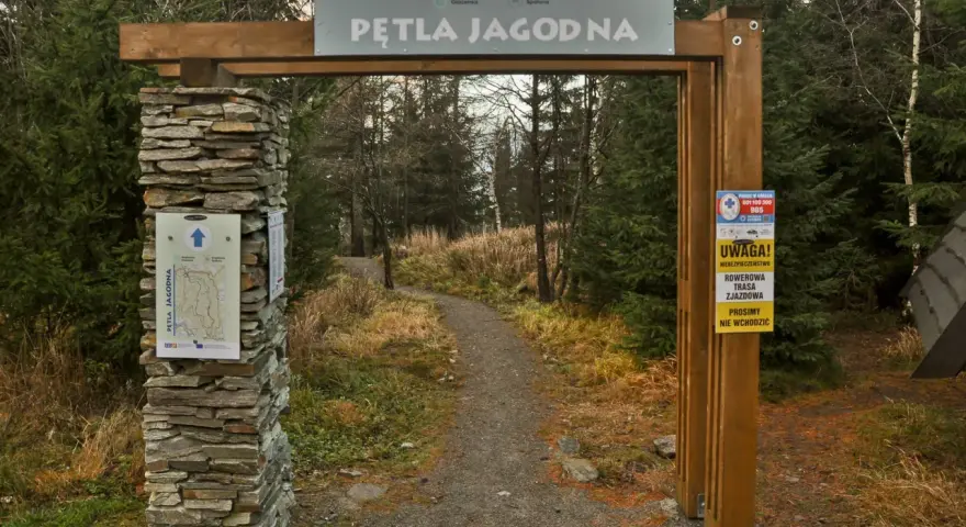 Kamienny filar i drewniana brama wita na Pętli Jagodnej. Ścieżka w lesie zaprasza do wędrówki.