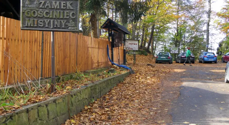 Tablica z napisem "Zamek GOSCINIEC MISYJNY" przy drewnianym płocie. Jesienny parking z samochodami i opadłymi liśćmi.