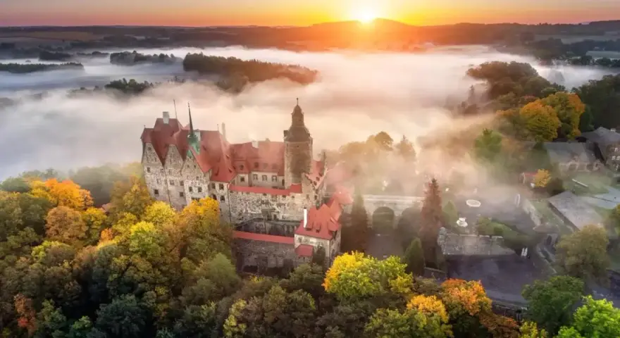 Zamek Książ otoczony jesiennymi drzewami, wyłaniający się z porannej mgły. Słońce wschodzi nad wzgórzami, malując niebo na pomarańczowo.