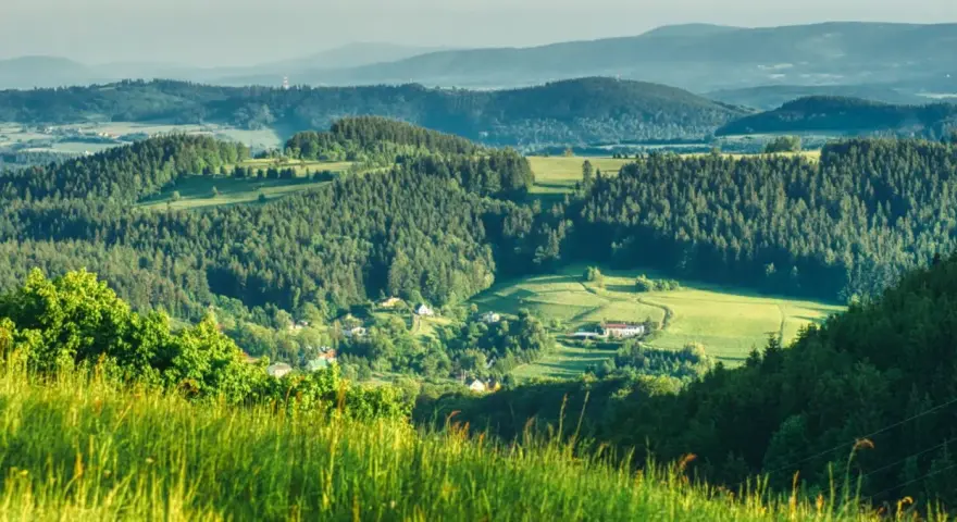 Zielone wzgórza i lasy Gór Sowich, z widocznymi domami i polami. Idealne miejsce na odkrywanie atrakcji i odpoczynek na łonie natury.